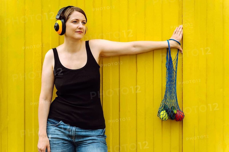 Cheerful woman with fresh veggies in mesh tote, urban style on yellow, Woman wearing headphones leans against a bright yellow wall holding a net bag with vegetables, creating a relaxed summer atmosphere