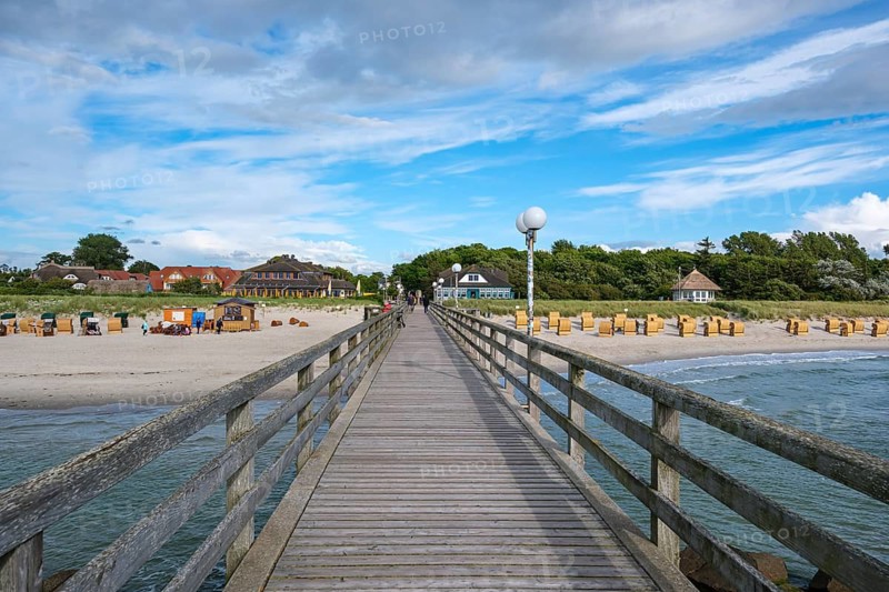 A long wooden walkway leads across the sea with a view of beach houses and beach chairs under a partly cloudy sky, Wustrow, Mecklenburg-Western Pomerania, Germany