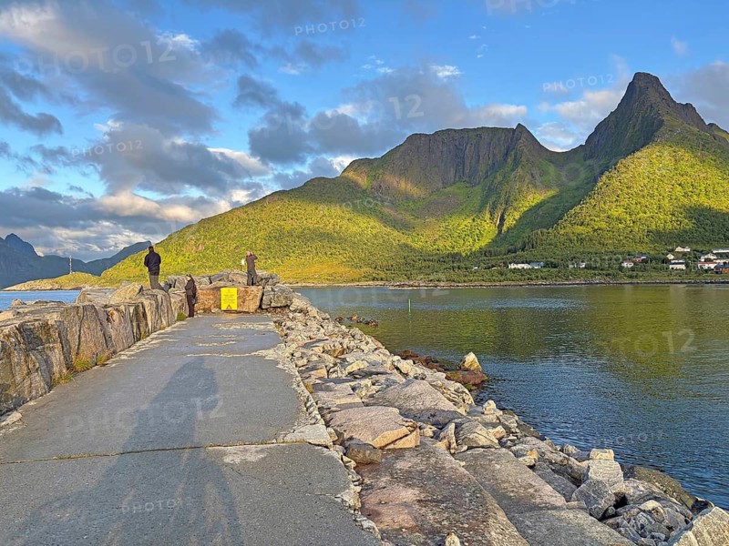 A picturesque view of high green mountains and a tranquil lake under blue skies, taken from a stony path