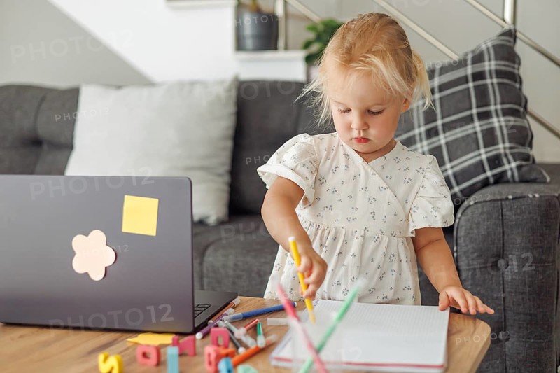 Young blond European girl drawing with colored pencils at home near laptop and sofa in living room
