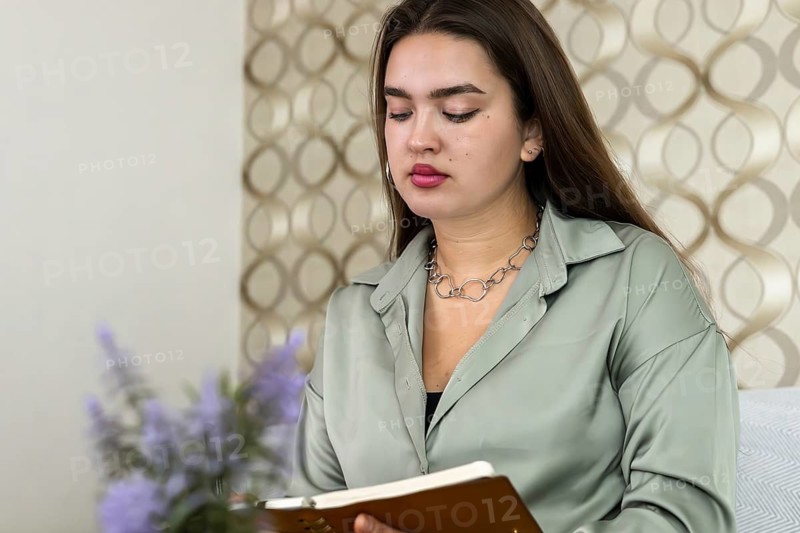 Young Hispanic woman with long brown hair writes in a notebook. She wears a green blouse and silver jewelry, sitting against a patterned wall