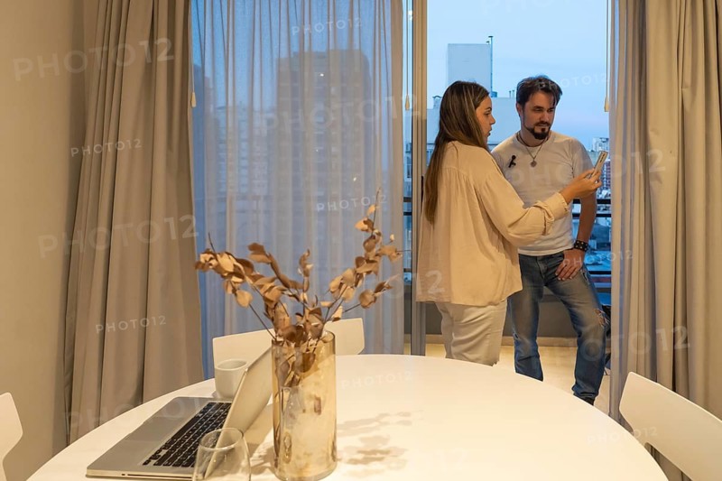 Woman and man standing close together reviewing video footage on smartphone screen near balcony doorway at dusk, city skyline in background