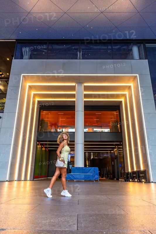 Young woman with curly hair walking confidently in a modern urban environment, embracing city life and fashion under the glow of architectural lighting at dusk
