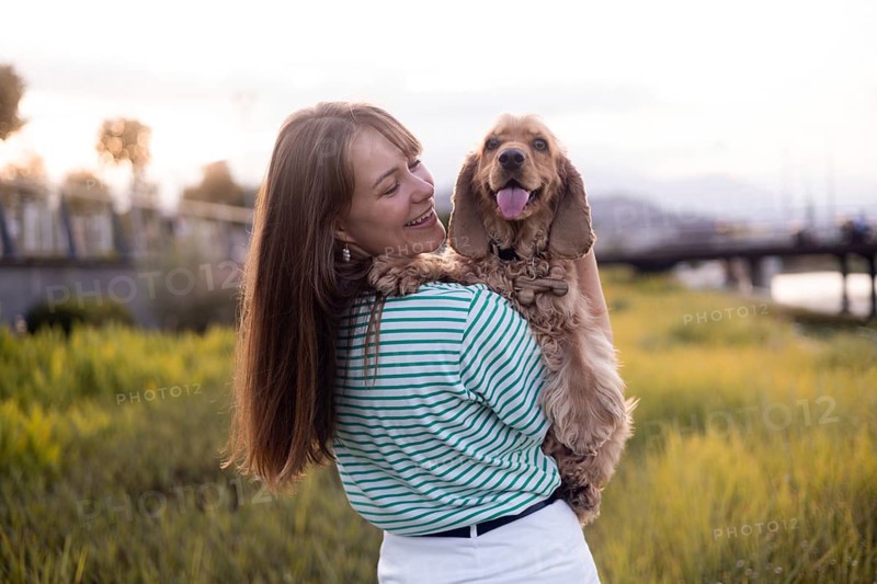 A happy woman embraces her dog Cocker Spaniel while standing in a sunset-lit field