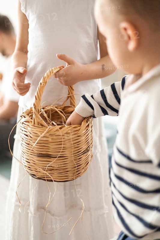 A young boy with short hair reaches into a woven basket held by a girl in a white dress. The scene captures a warm, family atmosphere at home. Easter eggs