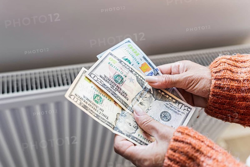 Woman holding US dollar banknotes near home radiator during winter heating season. Concept of rising home heating costs, expensive utility bills, inflation, household budgeting, energy affordability a.