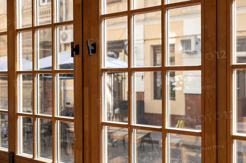 The warm ambiance of a cafe is highlighted by large wooden windows that frame a view of a lively outdoor terrace. Patrons enjoy their drinks at tables set up under umbrellas, while warm sunlight filters through the glass, creating a welcoming atmosphere. The thriving street hints at a vibrant community life just outside