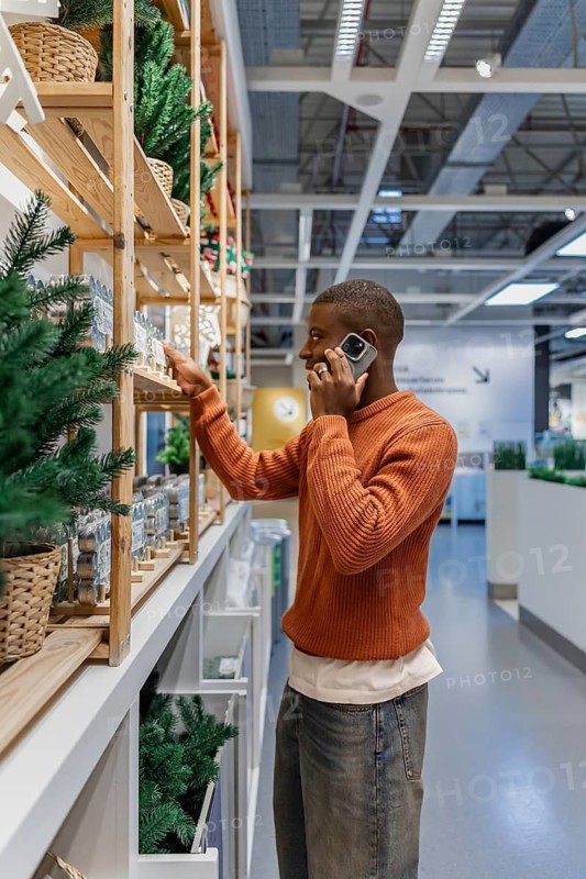 A man is standing in a store looking at home decor items on shelves. He is holding a phone to his ear while reaching for a product