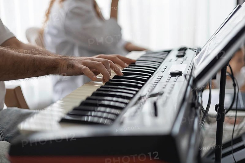 A close-up of a man's hand playing a keyboard in a cozy home setting. A woman is seated nearby, creating a warm atmosphere for a family concert