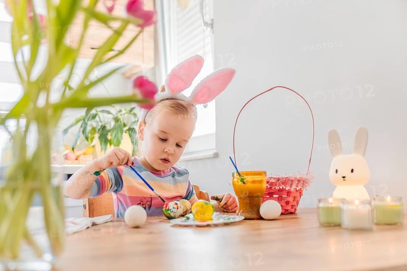 Toddler girl wearing pink bunny ears sits at table in kitchen decorated for Easter. Colorful eggs are on table. The room has bright light and festive items. The child looks cheerful and focused