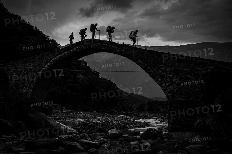 BENJA, ALBANIA - JULY 1, 2026: Black and white dramatic silhouette of hikers carrying backpacks walking across an ancient stone arch bridge in Albania