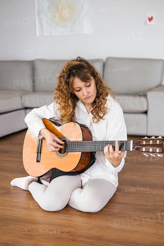 Woman practicing guitar on a hardwood floor, dressed in beige clothes