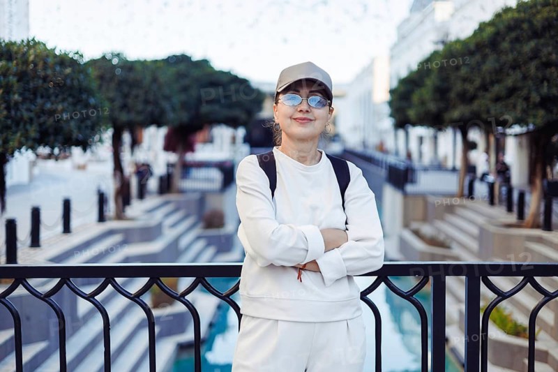 A woman wearing a cap and glasses stands with her arms crossed near a beautifully designed water feature. The surroundings are bright and lively, highlighting urban architecture