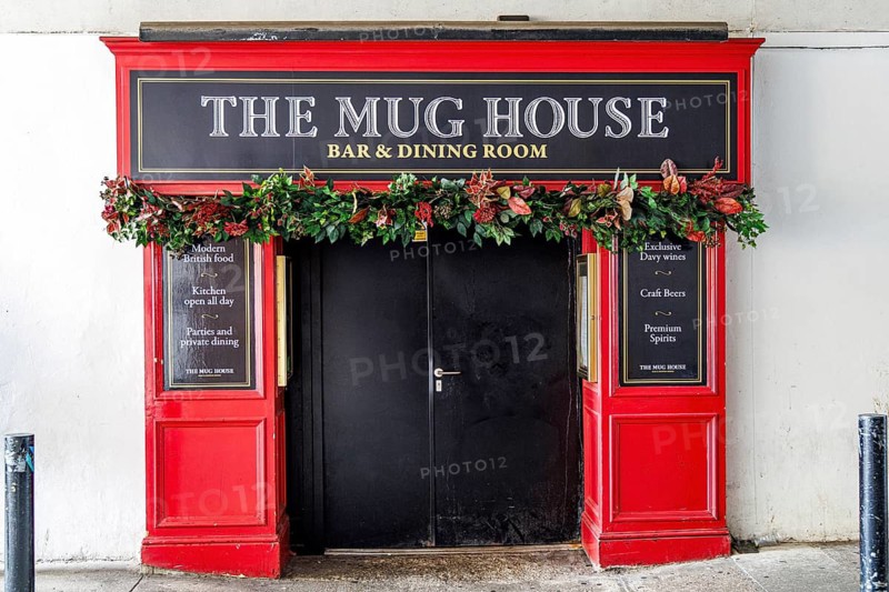 Floral garlands frame the dark doorway of The Mug House, a bar and dining room set beneath the brick arches near London Bridge, its bright red signage advertising modern British food, craft beers and premium spirits in Southwark, London, England, United Kingdom