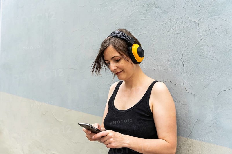 Blonde woman checking phone with headphones, urban lifestyle moment, Woman wearing headphones and looking at smartphone against a concrete wall