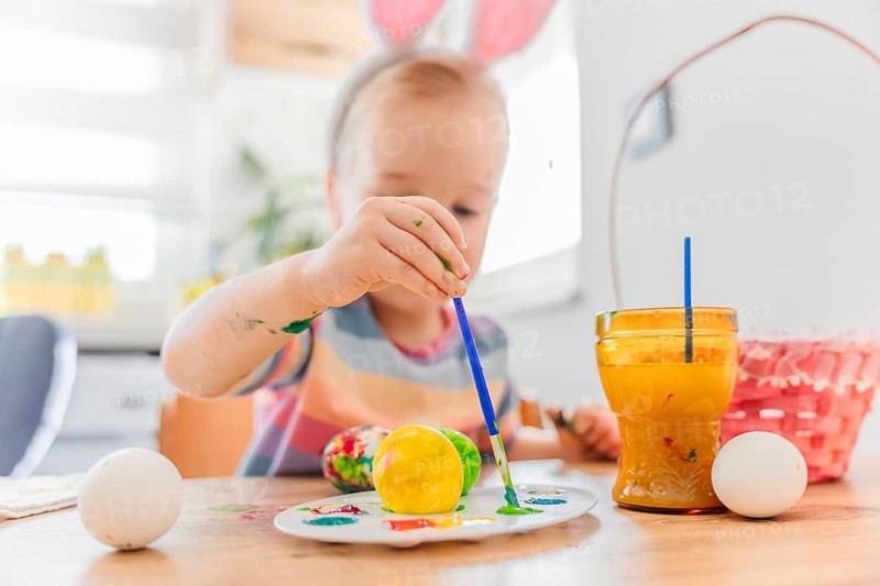 Toddler girl in dress wearing bunny ears painting Easter egg with brush, basket for eggs is near her. Spring holidays concept.