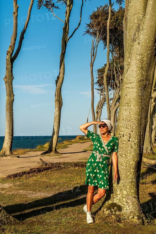 A woman (54) in a green tennis dress and white trainers is leaning casually against a tree, gazing into the distance, Gespensterwald, Nienhagen, Germany