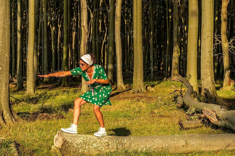 A woman (54) wearing a green tennis dress and white trainers is surfing on a tree, Ghost Forest, Nienhagen, Germany