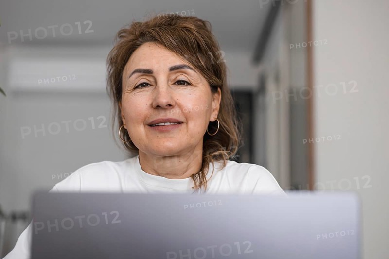 Portrait of mature woman sitting at home in the kitchen and using a laptop. Mid aged businesswoman working, studying from home and looking at camera and smiling