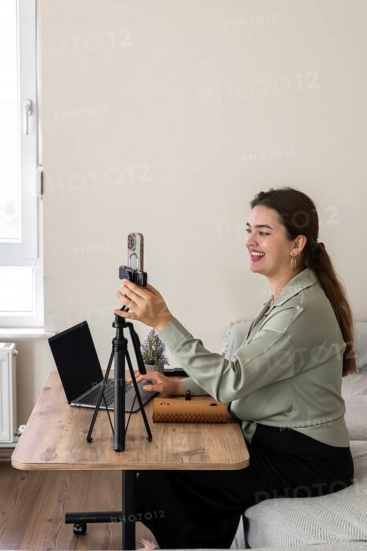 A young Caucasian woman with long brown hair sits at a wooden desk. She smiles while holding a microphone, with a laptop in front of her
