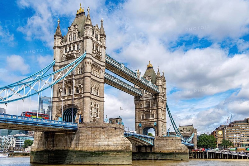 Viewed from the Thames footpath, the Gothic stone turrets of Tower Bridge frame its pale blue suspension walkway and lower bascule roadway, with a red London bus crossing between the towers on a bright summer afternoon, London, England, United Kingdom