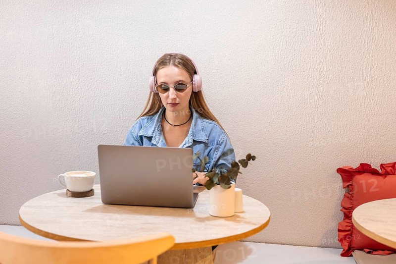 Young Caucasian woman with long brown hair wearing pink headphones, sitting at a table with a laptop and a cup of coffee, in a cozy indoor setting