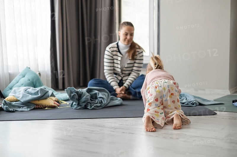 A young Caucasian woman sits on a mat, smiling at a small girl with blonde hair in a playful pose. The room is cozy with soft lighting and scattered blankets