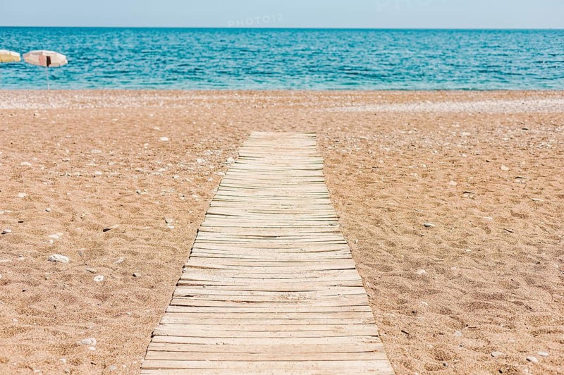 A wooden pathway leads to a calm beach with golden sand and turquoise water. The sky is clear, creating a serene coastal atmosphere