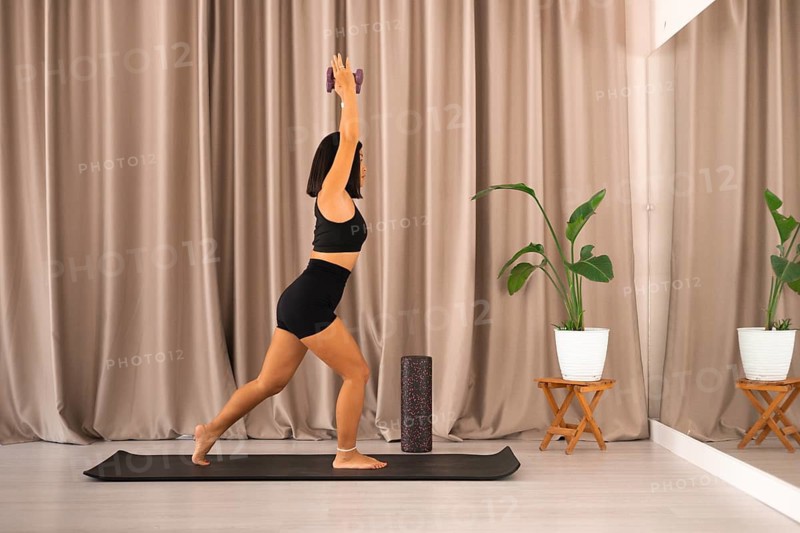 A woman stands on a yoga mat holding dumbbells, facing a mirror in an exercise studio