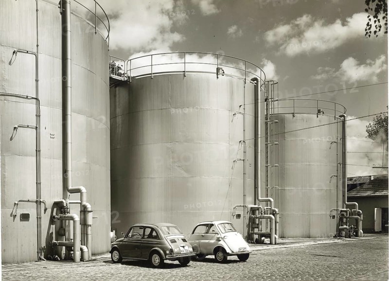 BMW Isetta, Fiat 500, small cars, gas tanks, large industrial tanks with two parked retro cars and cloudy sky above, around 1955
