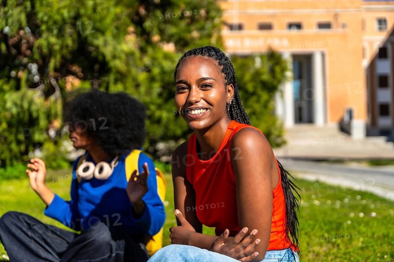 Smiling young african american woman sits on campus grass with a friend, enjoying a sunny break between classes, relaxed and engaged in conversation and study plans