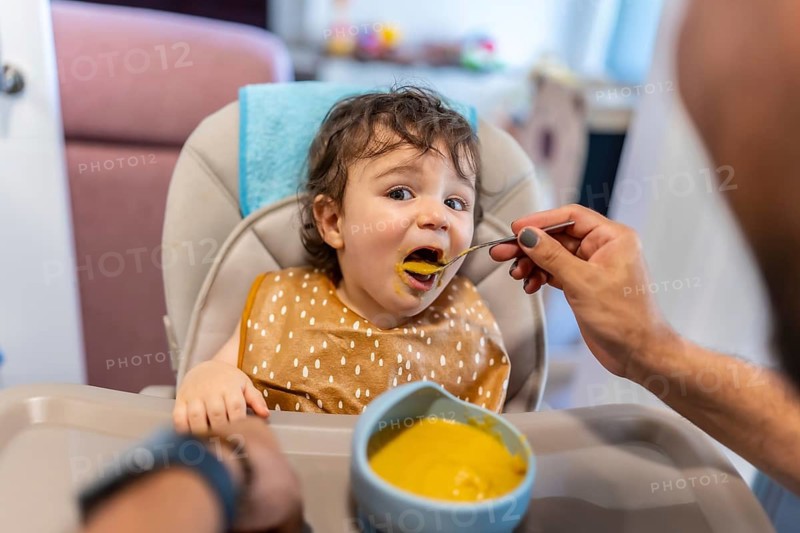 Baby boy in a high chair actively eating a spoonful of healthy vegetable puree, receiving food from his father's hand during a nurturing mealtime experience at home