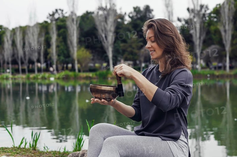 A young Caucasian woman with brown hair sits by a lake, holding a bowl. She wears a gray sweater and light gray pants. The background features trees and water
