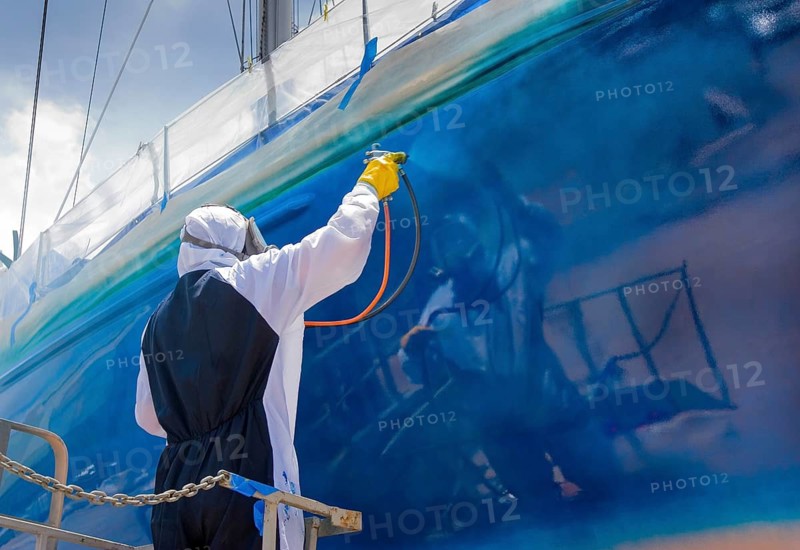 Worker spray paints the hull of a sailing yacht during professional marine maintenance, wearing protective clothing and equipment