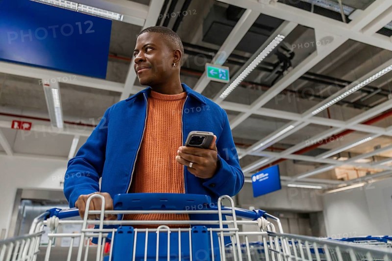 A person stands in a store with a shopping cart, holding a mobile phone. They are looking around while browsing the aisles during the day
