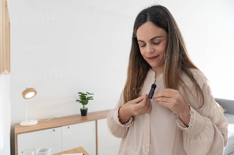 Close up of woman attaching small lavalier microphone to her blouse collar, preparing to record video content in bright home studio setup