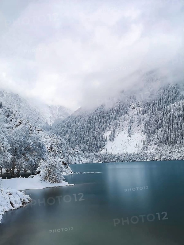 Low clouds over the turquoise water of the mountain lake in winter. Winter landscape, A serene winter landscape with snow-covered mountains and a calm lake under a misty sky, Lake Issyk, Kazakhstan