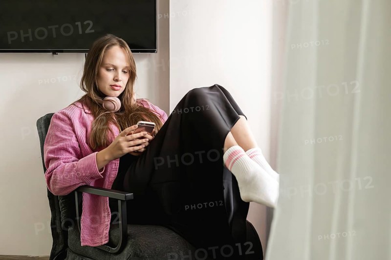 A young woman relaxes in a chair with her feet on the seat while using her mobile phone. She wears headphones and sits in a room with soft, natural light