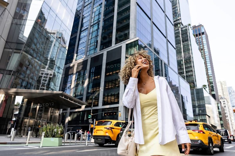 Young woman with curly hair smiling and talking on her mobile phone while walking on a bustling manhattan street with skyscrapers and yellow cabs in the background
