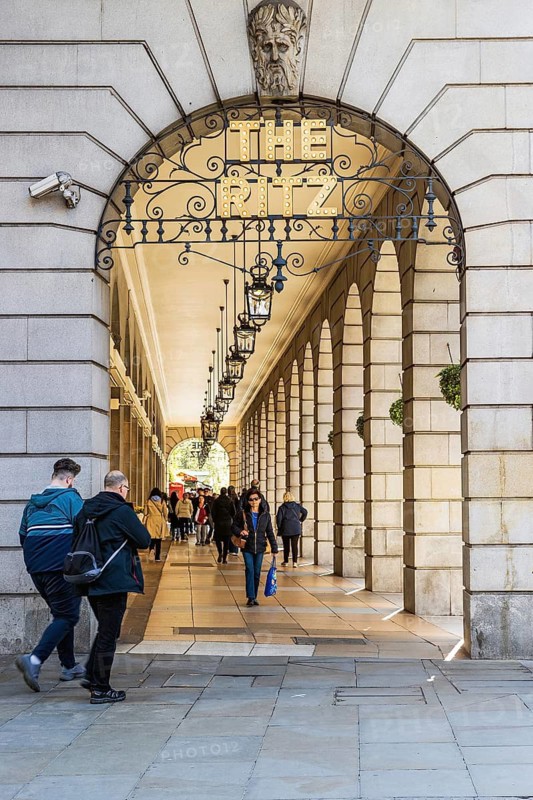 Pedestrians pass beneath the stone arcade and lanterns of The Ritz Hotel on Piccadilly, London, England, United Kingdom