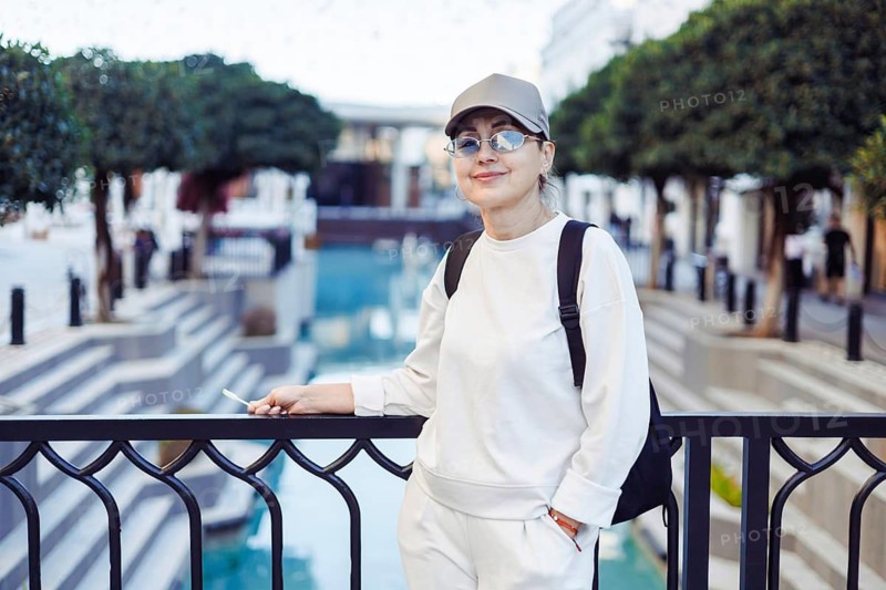Middle-aged woman stands casually by a railing, smiling while overlooking a serene water feature in a contemporary urban environment. The scene captures a tranquil moment amid bustling city life