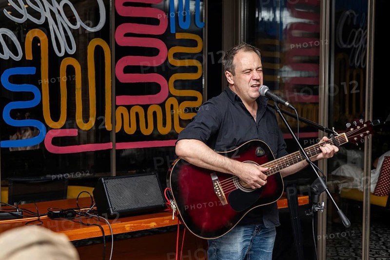 A middle-aged Caucasian man with short brown hair sings into a microphone while playing an acoustic guitar. Colorful background with abstract patterns