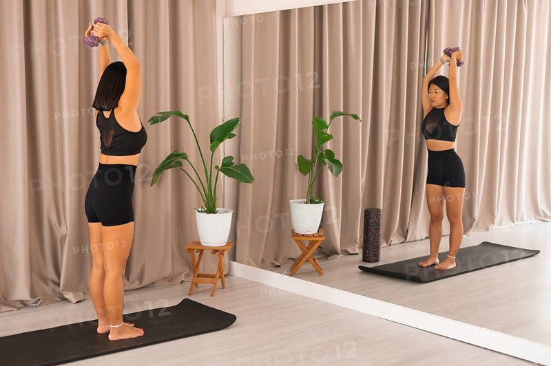 A woman extending arms with dumbbells above her head in front of a mirror