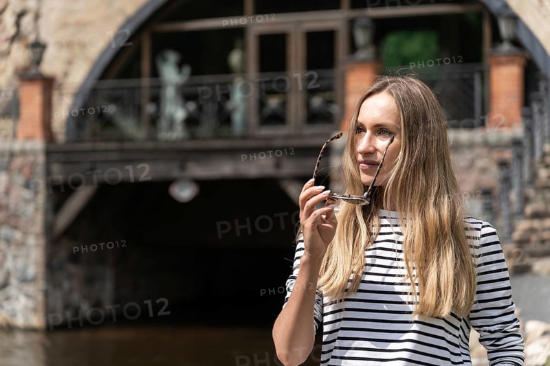 Long-haired woman takes off her sunglasses looking away, Woman holding sunglasses near a bridge with water flowing underneath, wearing a striped shirt in an urban setting, Belmontas Park, Vilnius, Lithuania