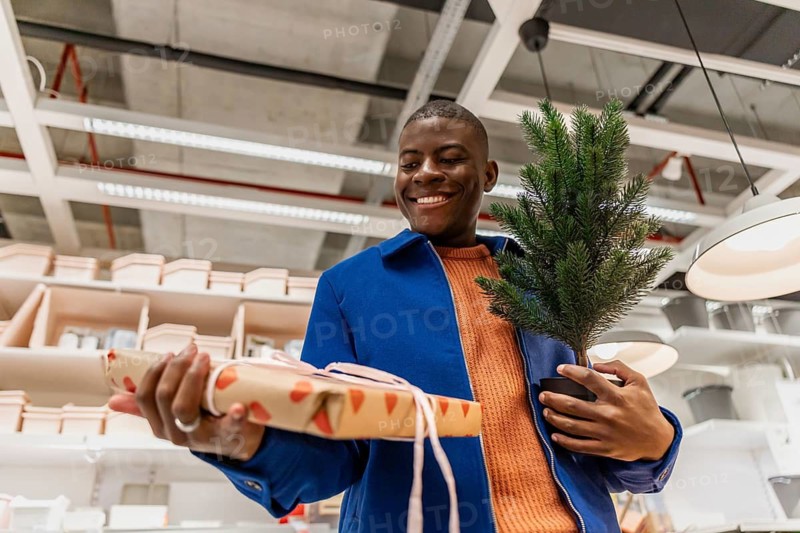 A person stands in a store with a smile, holding a wrapped gift in one hand and a small green plant in the other. The background shows shelves with various items