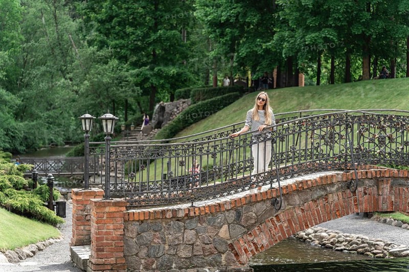 A woman stands on an old stone bridge with a forged fence in the park in the summer, Woman standing on a stone bridge with metal railings in a lush green park, Belmontas Park, Vilnius, Lithuania