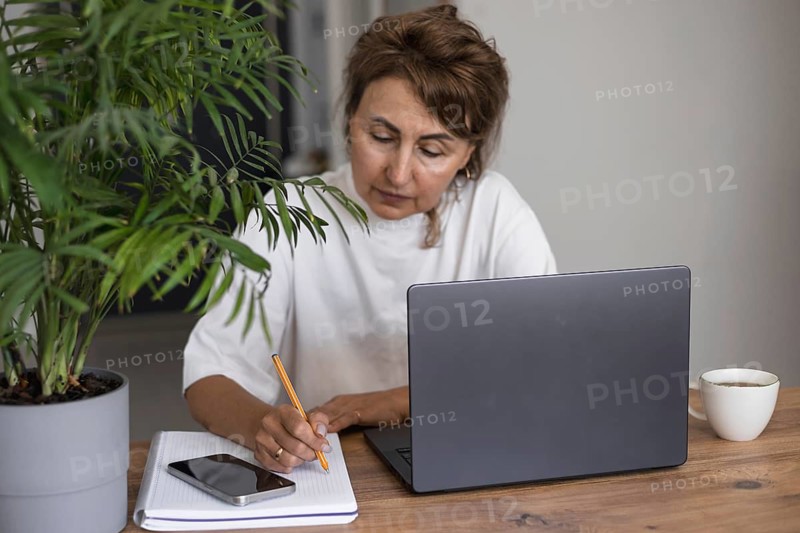 Serious pretty mature woman in white outfit studying at laptop at home, writing notes in notebook, watching online training webinar, professional course, listening to audio lecture