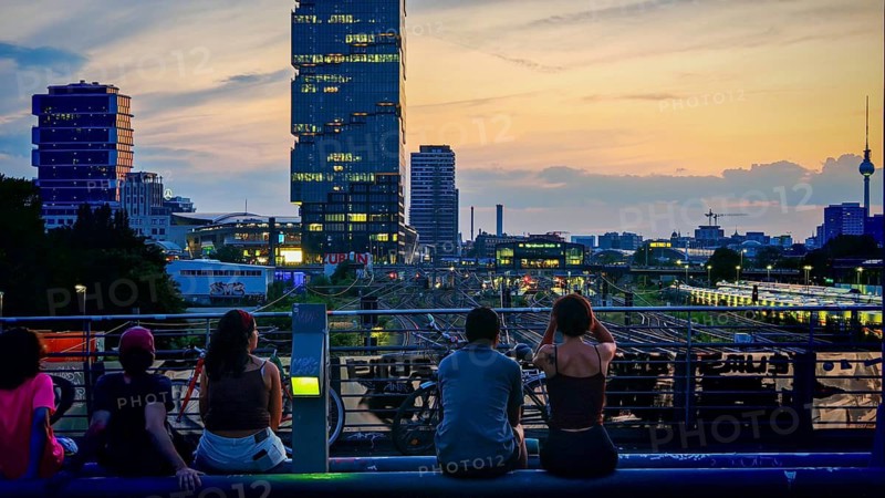 Several people enjoy the sunset on the Modersohn Bridge with a view over train tracks, television tower and a modern cityscape, Berlin-Friedrichshain, Berlin