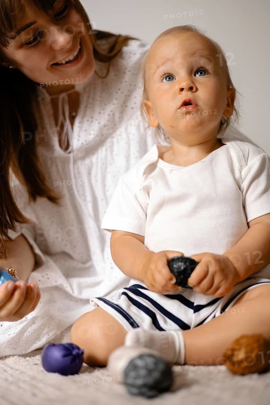 Baby holding a toy, gazing upward next to a smiling mother