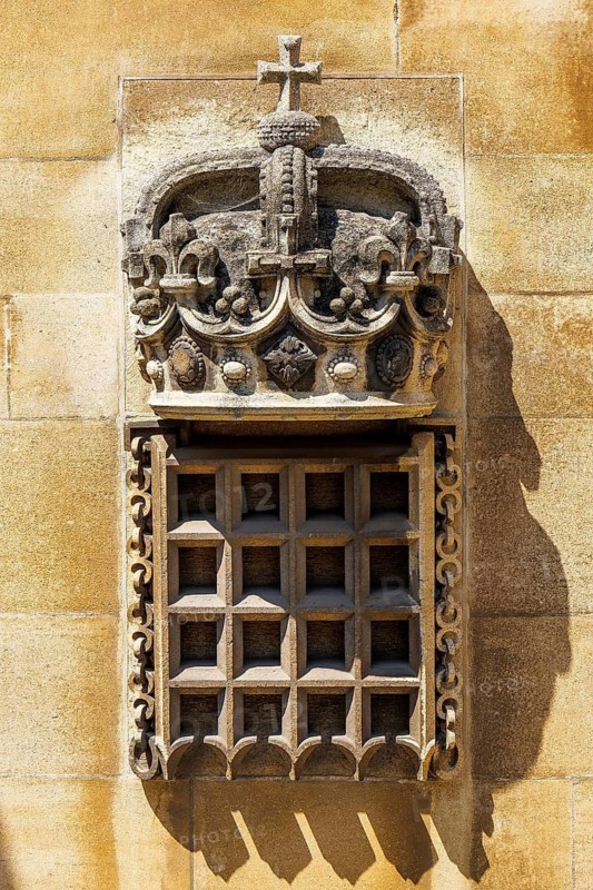 A stone carving of a crowned portcullis hangs on chains above a latticed grille set into a sandstone wall at Windsor Castle, the portcullis being an ancient heraldic badge symbolising royal authority and the castle's fortified origins, Windsor, England, United Kingdom